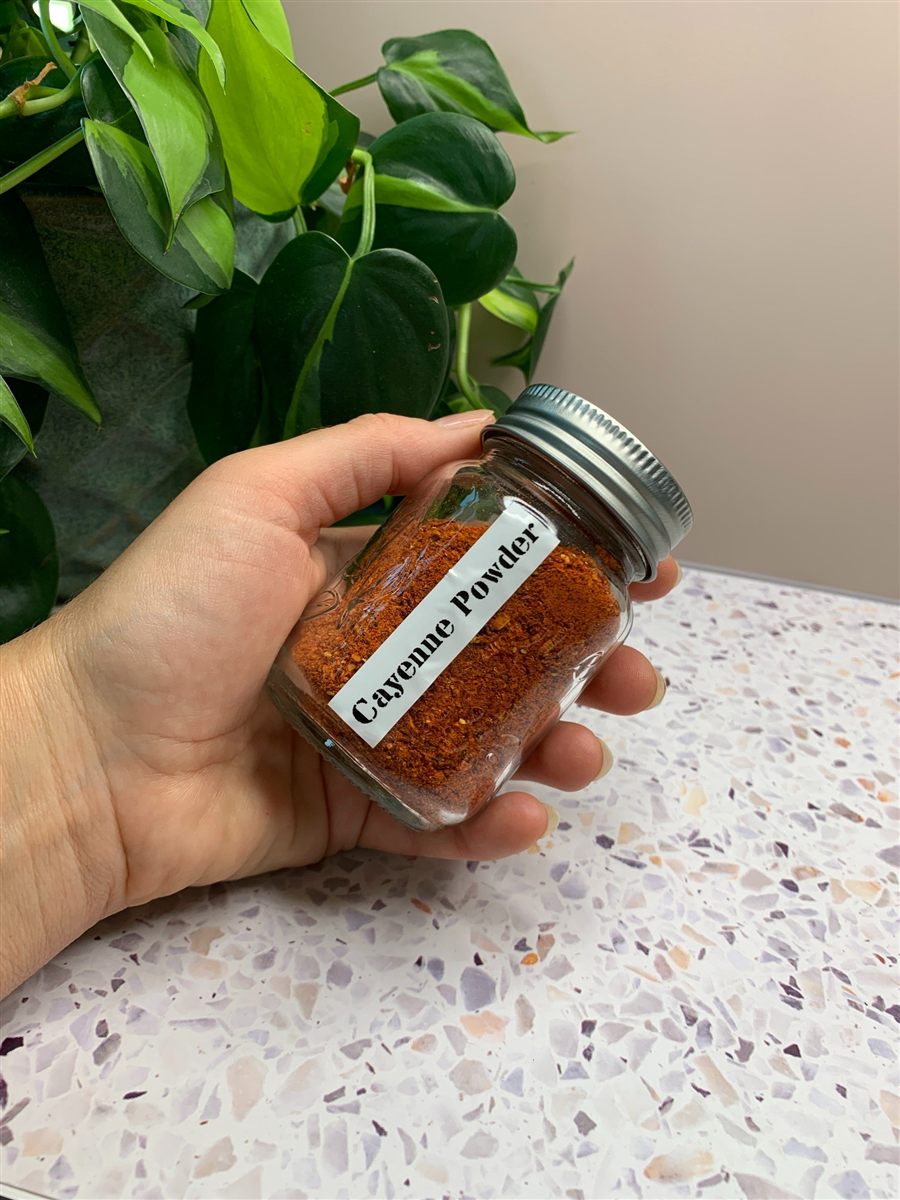 A hand holding a small jar labeled Cayenne Powder, in front of a potted plant.
