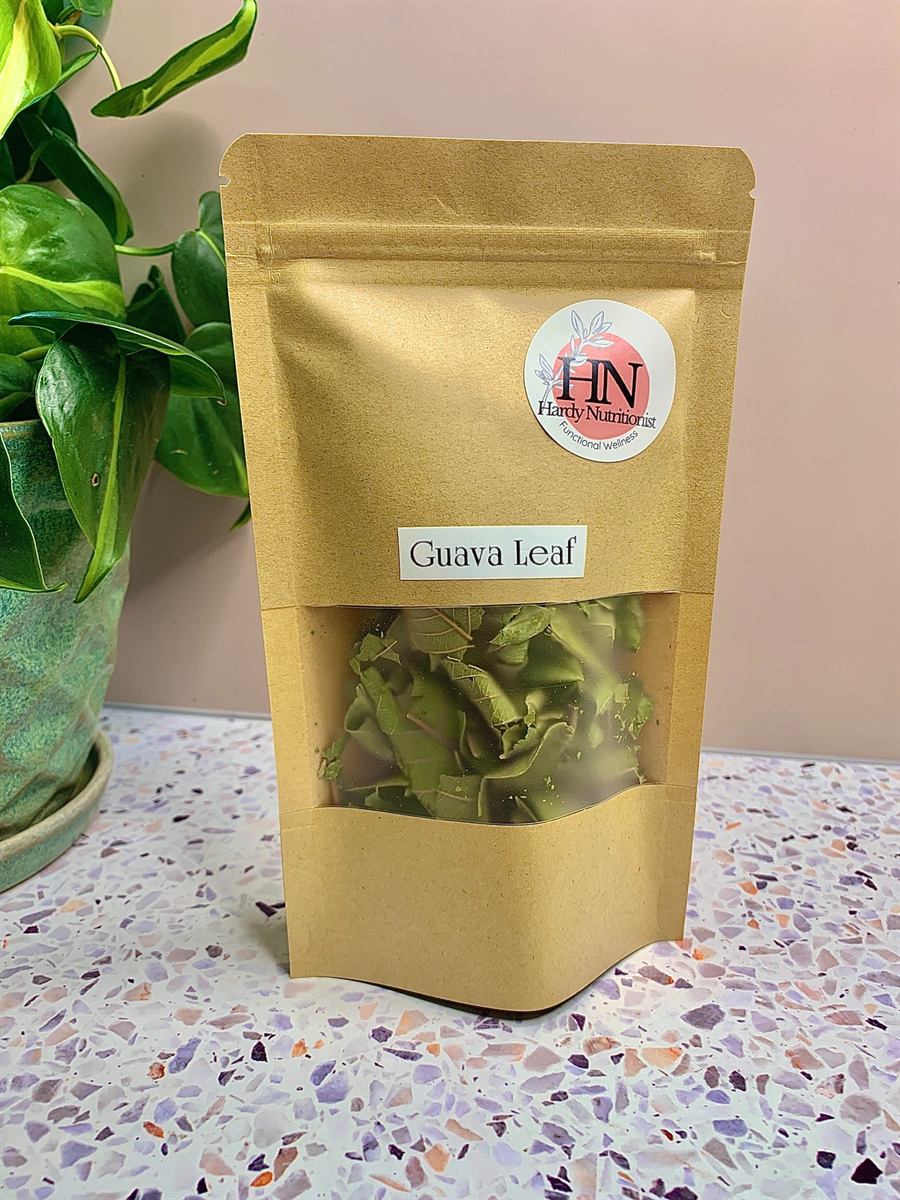 A kraft paper pouch labelled Guava Leaf, carrying a round Hardy Nutritionist sticker, its clear window showing dried guava leaf pieces. A potted pothos stands beside it on a terrazzo surface.