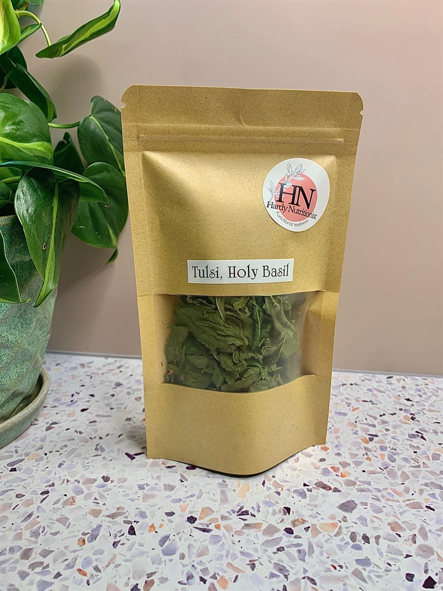 A kraft paper pouch labelled Tulsi, Holy Basil, carrying a round Hardy Nutritionist sticker, its clear window showing whole dried tulsi leaves. A potted pothos stands beside it on a terrazzo surface.